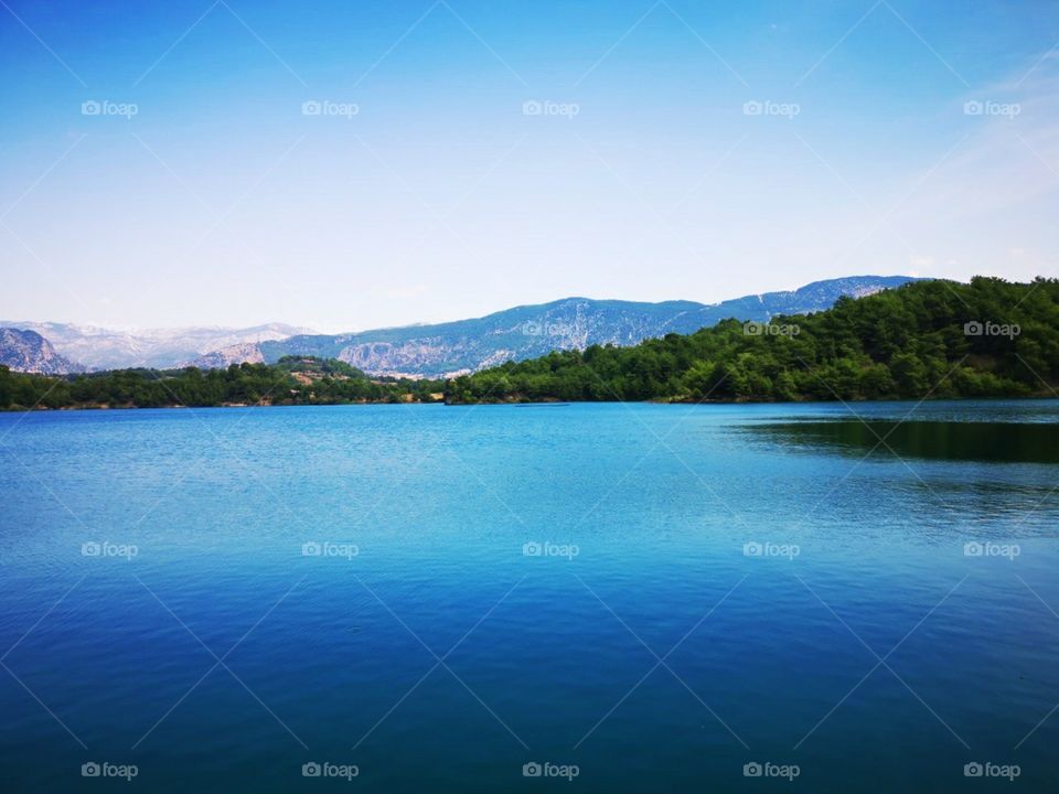 A beautiful photo of a turquoise blue lake with reflection of shadow of a tree. In the background are mountains and nature. Made on sunny day.