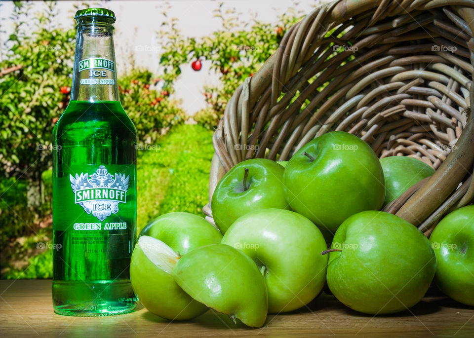 green apple flavor bottled vodka cooler on wood table next to a basket of green apples spilling across the table with an orchard in the background