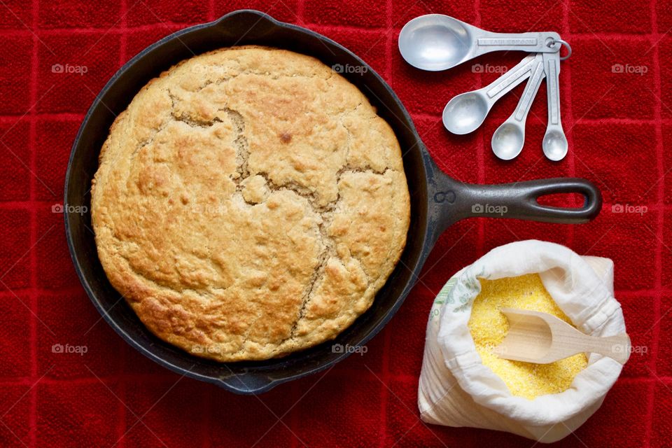 Flat lay of sourdough cornbread in a cast iron skillet, cornmeal and wooden scoop in a flour sack, vintage measuring spoons on a red kitchen towel