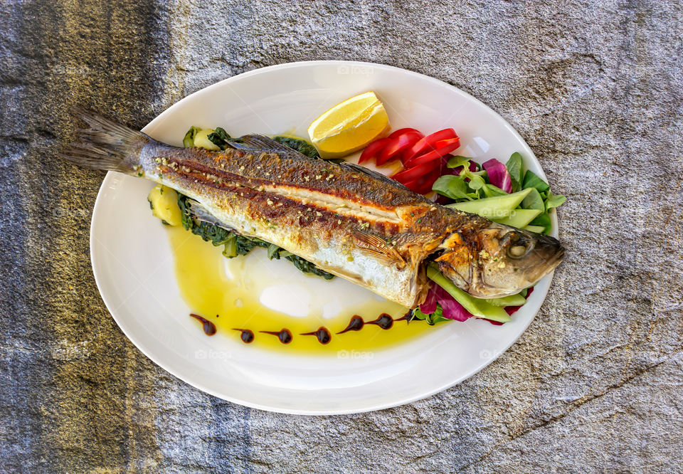 The concept of a healthy balanced Mediterranean diet. Croatian, Dalmatian cuisine: sea bass baked with spinach and vegetables. Grill fish with herb, lemon on white plate on a natural stone background