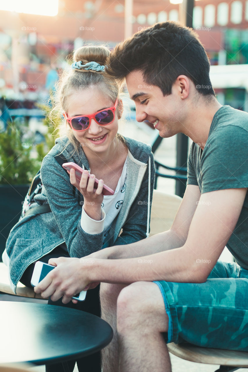 Couple of friends, teenage girl and boy, having fun using smartphones sitting in center of town, spending time together