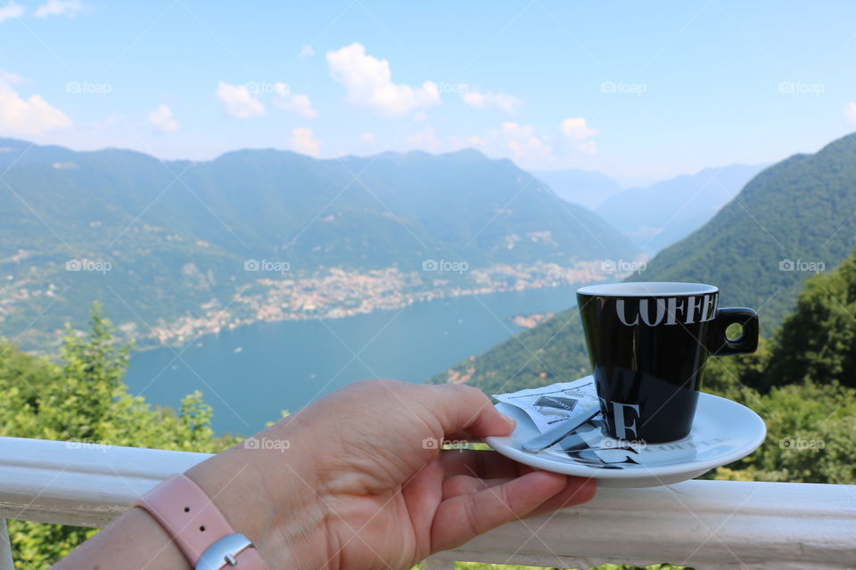 Hand holding coffee cup over the fence of a balcony overlooking the beautiful scenery of lake surrounded by mountains, on a bright summer day