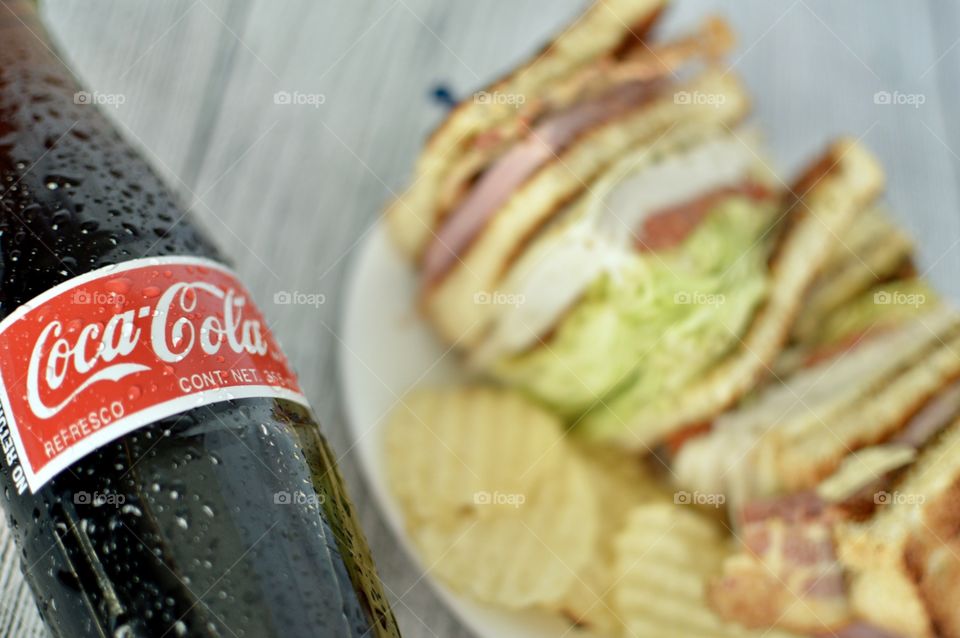 A can and a Bottle of Coca Cola with a club sandwich on a white rustic background 