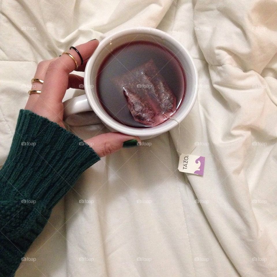 Arm wearing green sweater with rings on fingers holding a hot mug of tazo tea