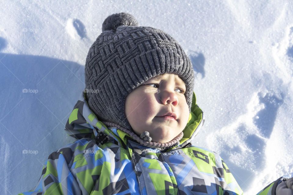 A child with a serious expression on his face in winter clothes jackets, pants, hat and boots in winter on the white snow on the street and in the park in nature plays winter fun.