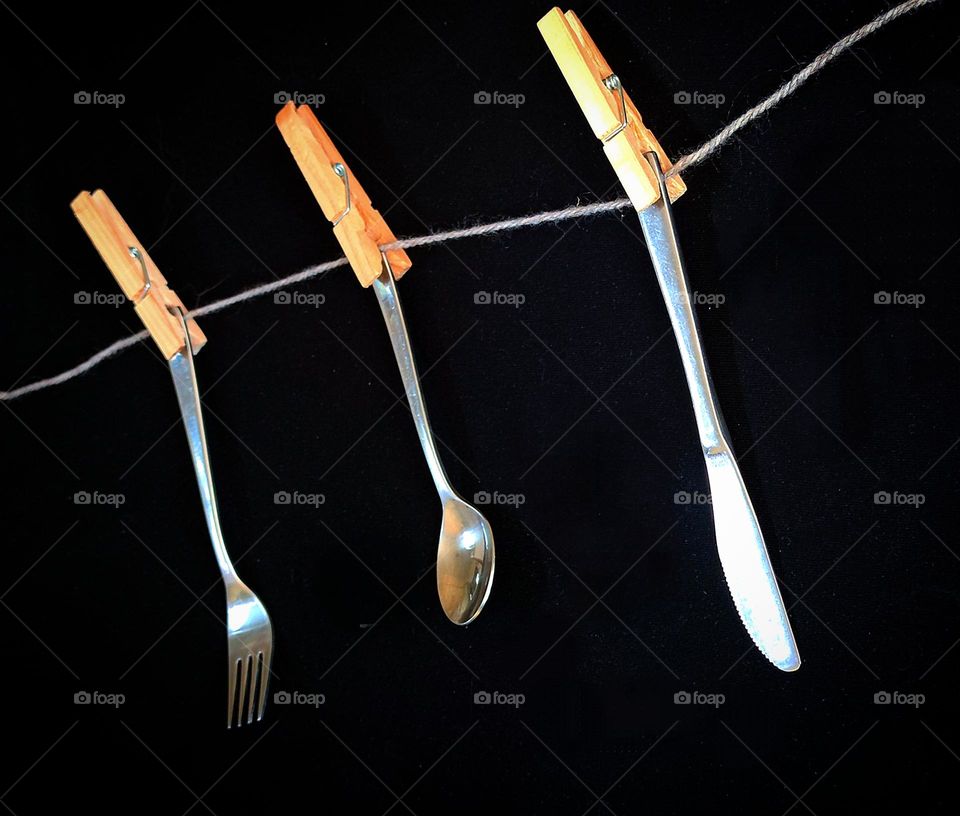 Metal. Wood.  On a gray rope hang metal cutlery attached with wooden clothespins: a fork, a spoon and a knife.  Black background.Combination of warm wood and cold metal