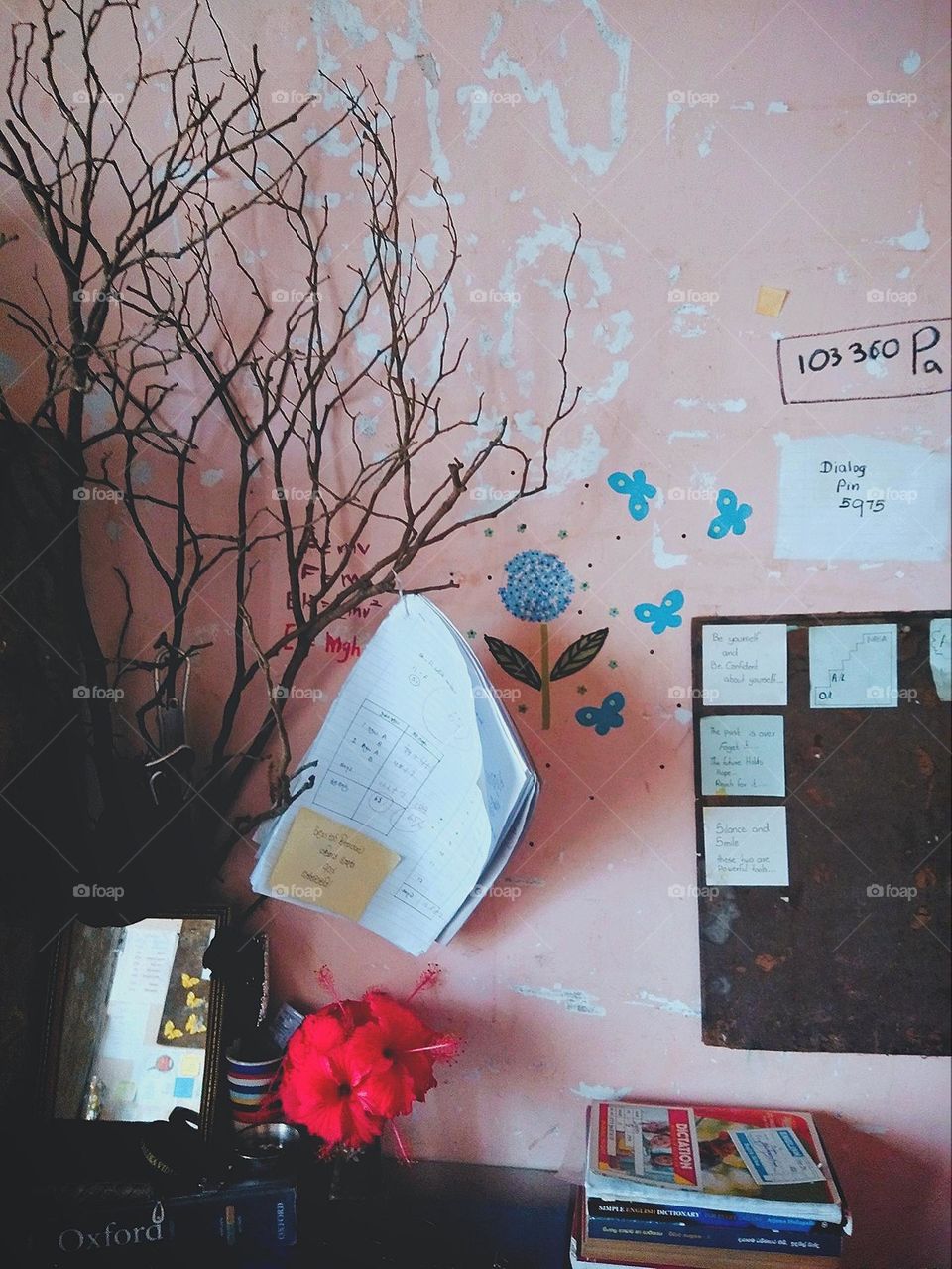 This is a capture of a study table. There is a another level of aesthetic to see.there is a wallpaper , wall art, flower vase , a mirror , a brown colour tree branch and there is a paper hanging on branch.And also there are some books to be seen.