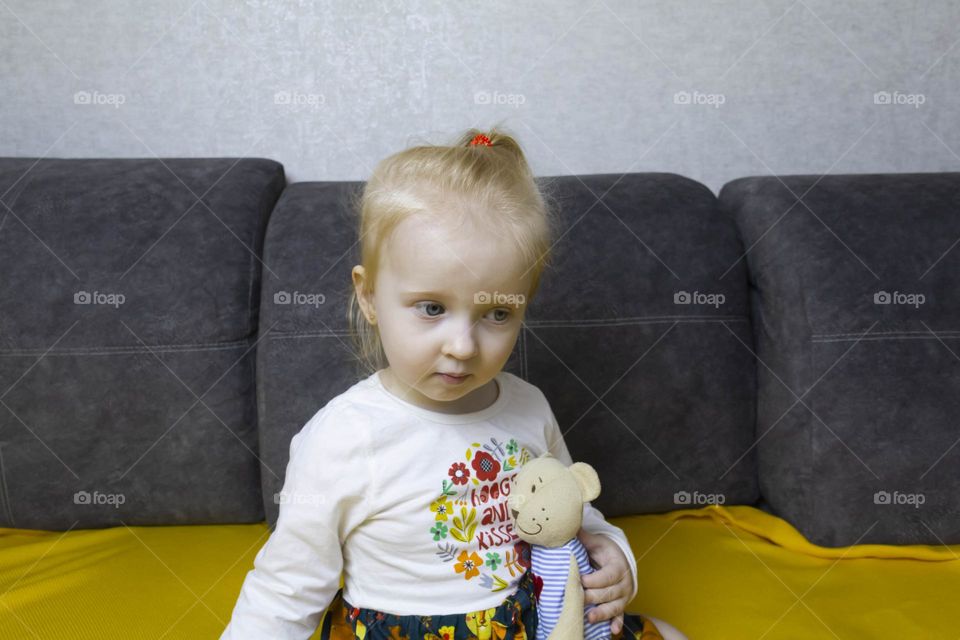 A small, red-haired girl is laughing at home on a gray sofa with purple pillows and a yellow background