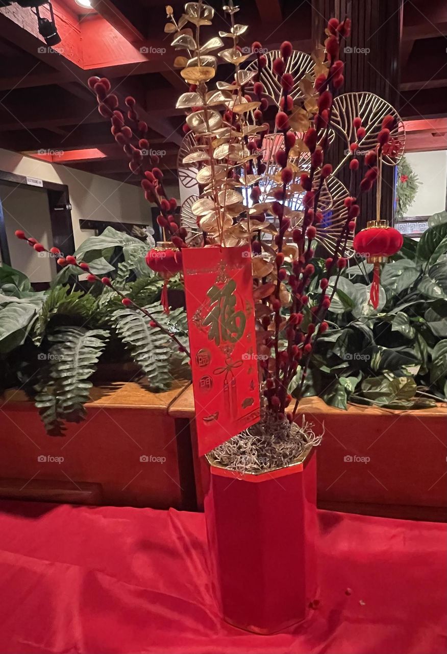 The centerpiece features a vibrant display on a rich red tablecloth, symbolizing prosperity and good fortune for the Chinese New Year. Hanging from the ornament is a red envelope (hongbao), symbolizing blessings and well-wishes for the upcoming year.