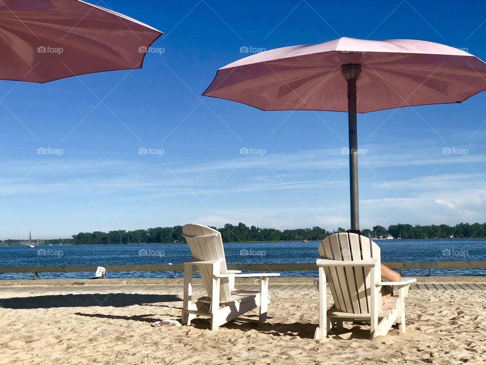 Pink umbrellas and white muskoka chairs on sand beach close to the blue water of the lake