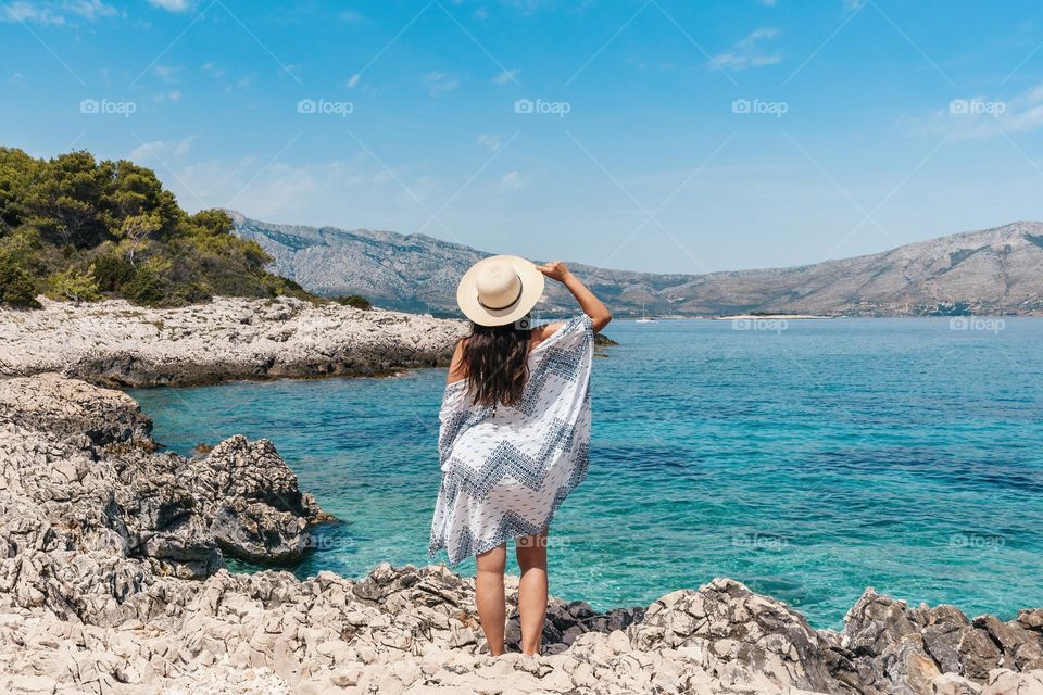 Rear view of young woman wearing beach clothes and sun hat standing on rocky sea shore on a sunny day in summer