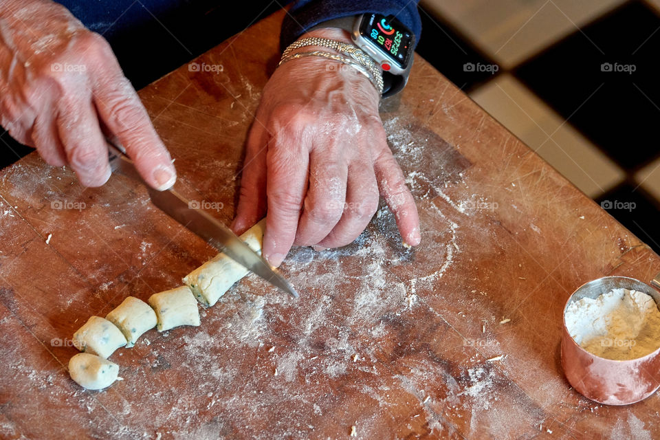Shot of tech-savvy old woman’s hands making and cutting fresh gnocchi in her kitchen on a floured butcher block. Shows skill, fresh ingredients, finely-honed skills, and cooking with love. Contrasts tradition with modern tech.