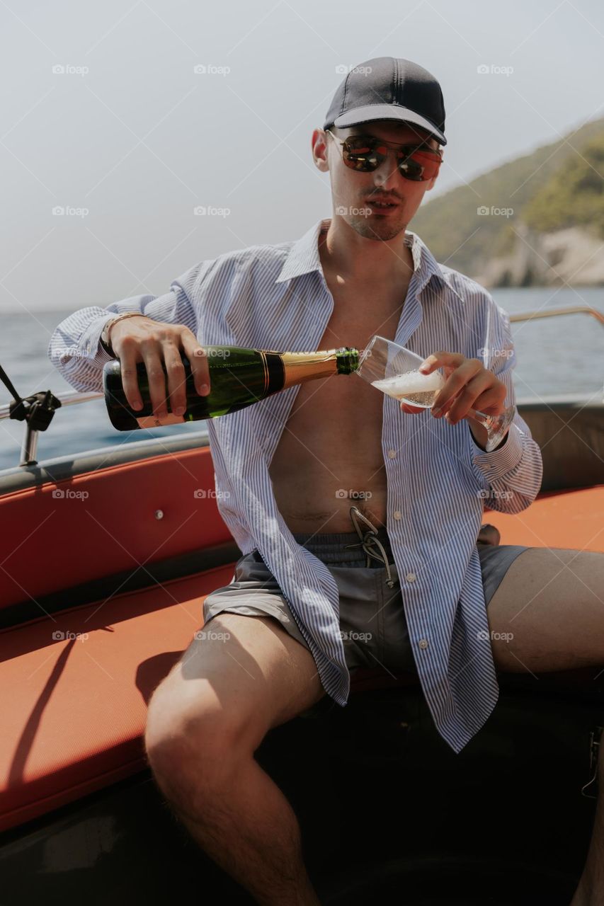 Portrait of one handsome young Caucasian man in a cap and sunglasses pouring champagne from a bottle into a glass, sitting in a boat on the sea on a sunny summer day, close-up side view.