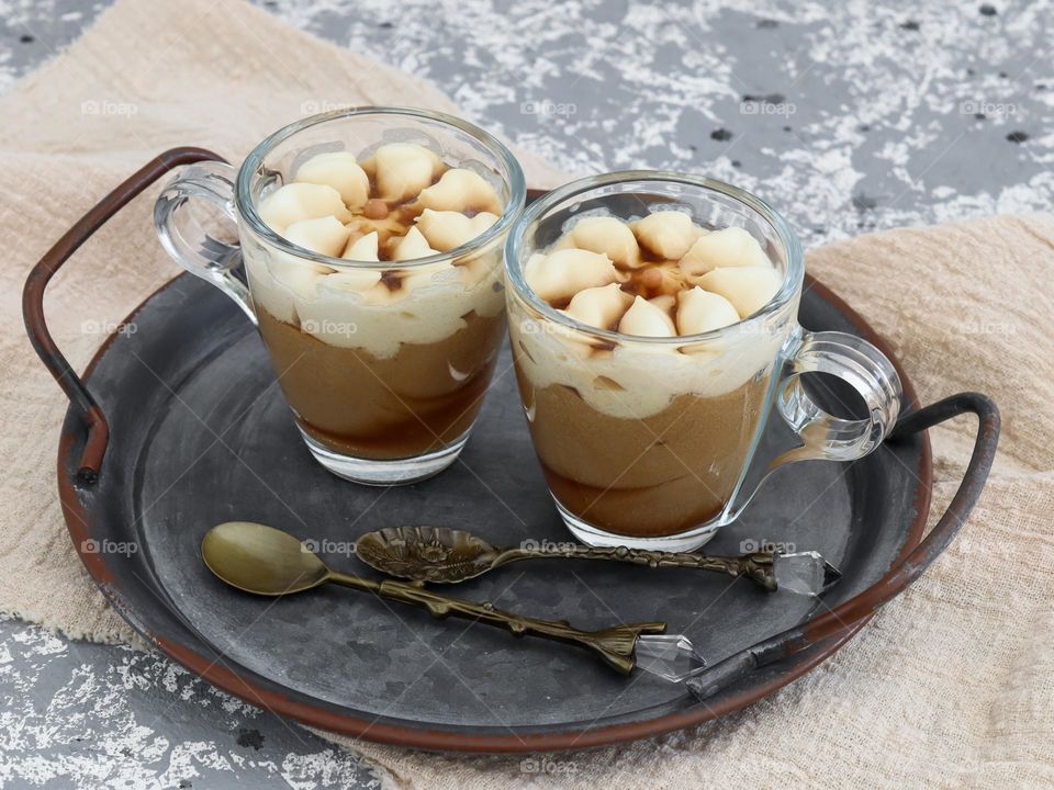 Two small glass transparent mugs with hot coffee and whipped cream on a vintage coogle platter with handles and retro dessert boats with a kitchen napkin lie on a gray-white stone background, close-up side view. The concept of hot drinks, sweet desse