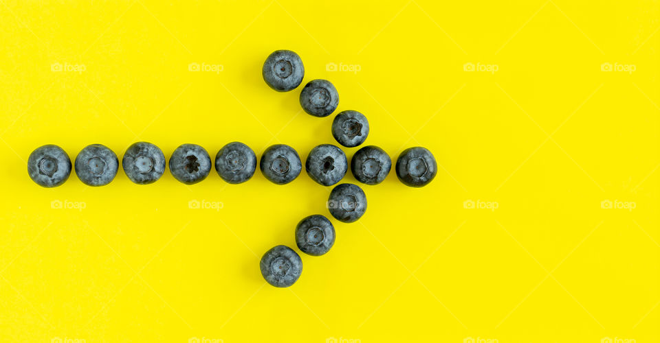 Organic blueberries arranged in the shape of arrow on yellow background, flatlay