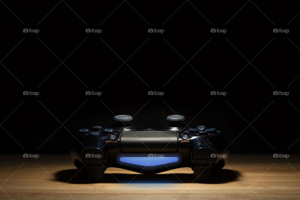 A portrait of a black playstation 4 controller being hit by a spotlight 1nd surrounded by darkness. the device is turned on and the light is blue.