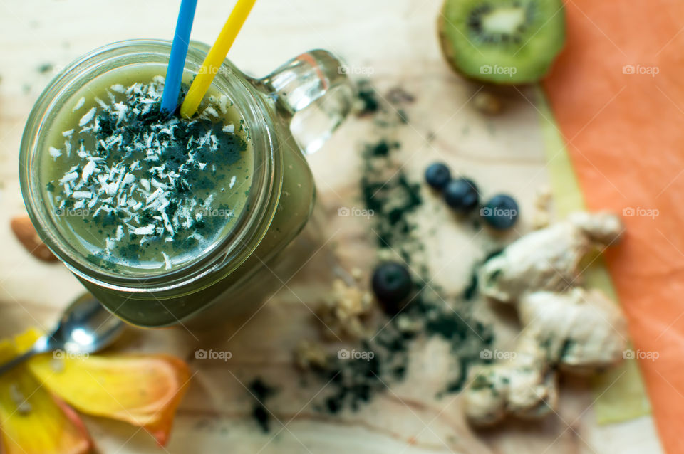 Making a sweet green fruit smoothie high angle view of spirulina protein powder and coconut in sweet green smoothie on table in glass jar with drinking straws with kiwi, ginger root and almonds in background
