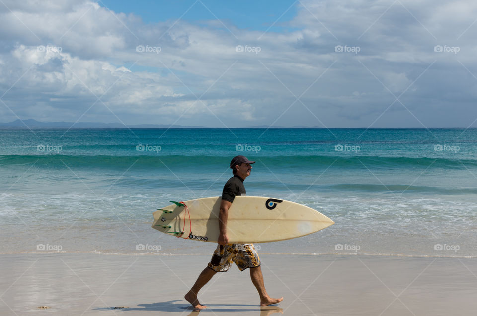 Surfer walks in the beach holding his surfboard. Photo taken in Byron Bay, Australia.