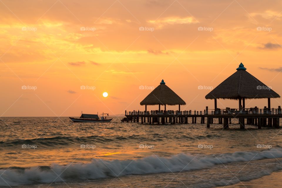 Orange Sunset in Cambodia. Orange sunset taken at the seashore in Cambodia. Couple straw huts on pier. One small boat. Waves crashing to the shore