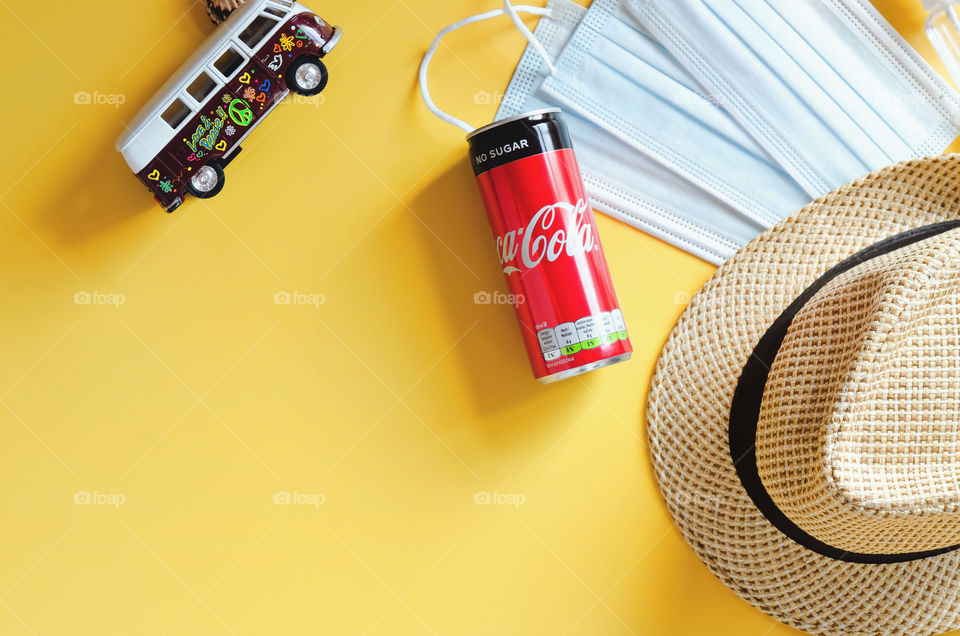 Travel under Covid-19 and new normal concept. Top view of medical face mask, can of Coca-Cola, phone and beach hat on yellow background. Creative ideas of prevent Coronavirus.