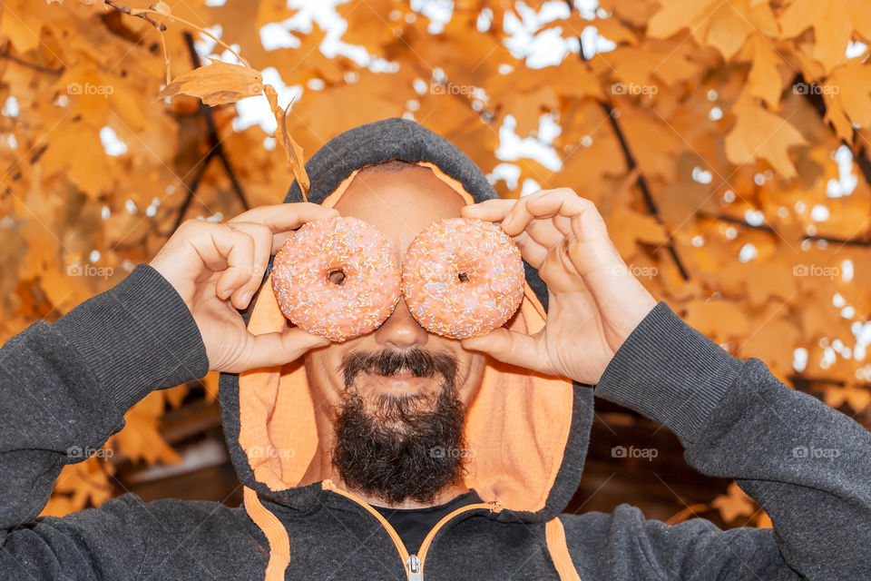 Poryrait of happy man in casual clothes have fun with two doughnuts on autumnal background.