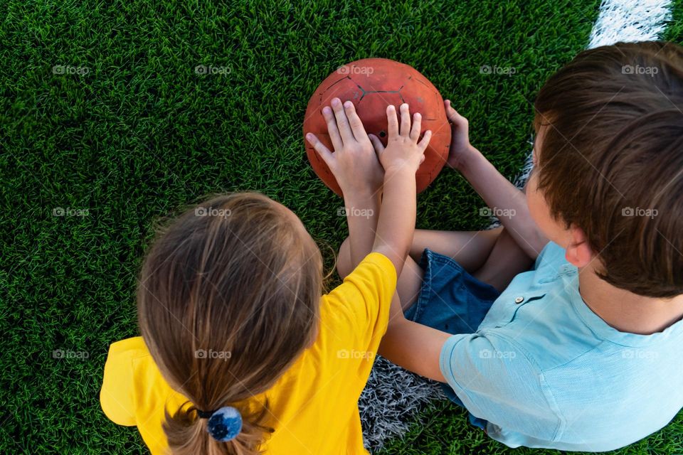 Back top view of anonymous kids sitting on lawn and holding ball while spending time