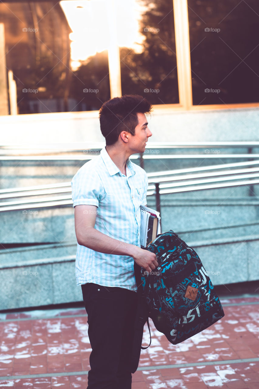 Student putting a notebook to a backpack. Boy standing at front of university building. Young boy wearing blue shirt and dark jeans