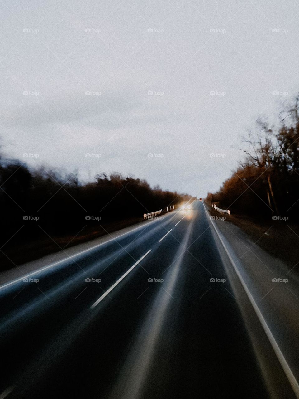 Car with lights on rides on the highway. Car road trip. Gray asphalt highway runs the horizon, trees along the way. Evening sunset sky. Car rides on the road. View from the car window. Eastern Europe, Ukraine