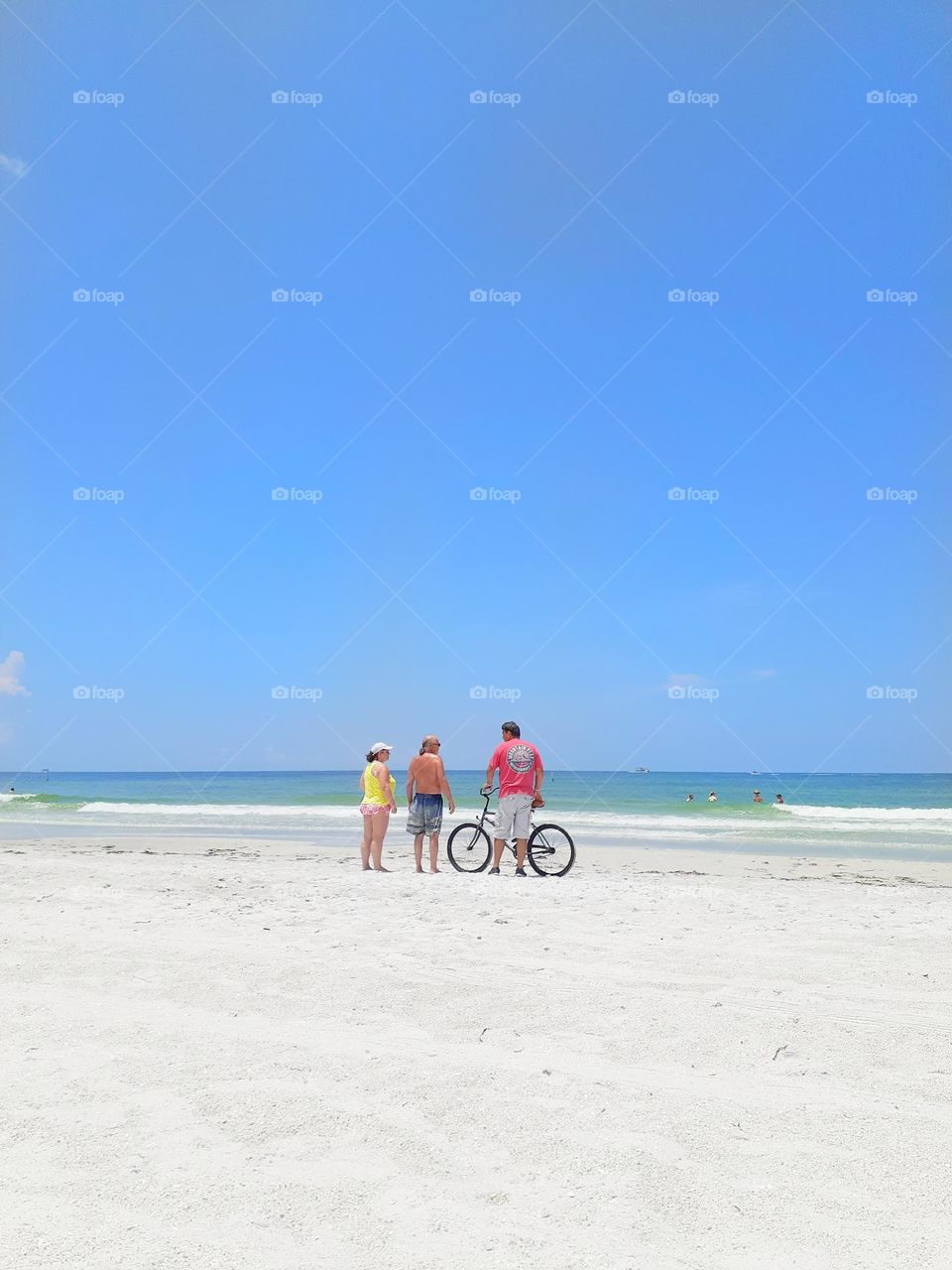 Three friends, two men and a woman stand on Indian Rocks Beach talking. One man is holding a bicycle. It is a beautiful day.