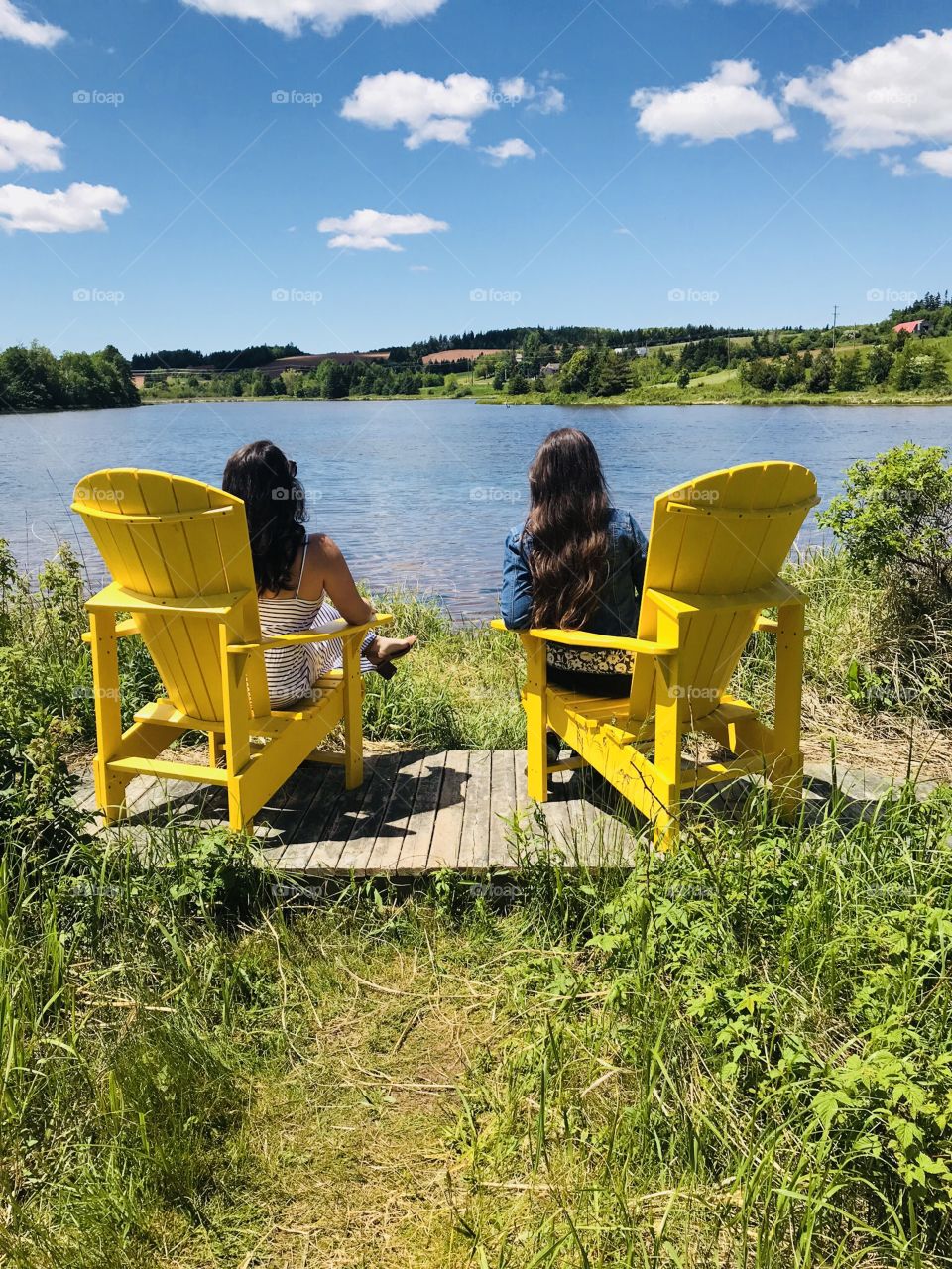 A happy picture of two fashionably dresses cousins enjoying the beautiful view of Prince Edward Island, Canada in bright yellow chairs. Surrounded by lush vegetation and bright blue skies, the whole scene captures a perfect moment of summer.