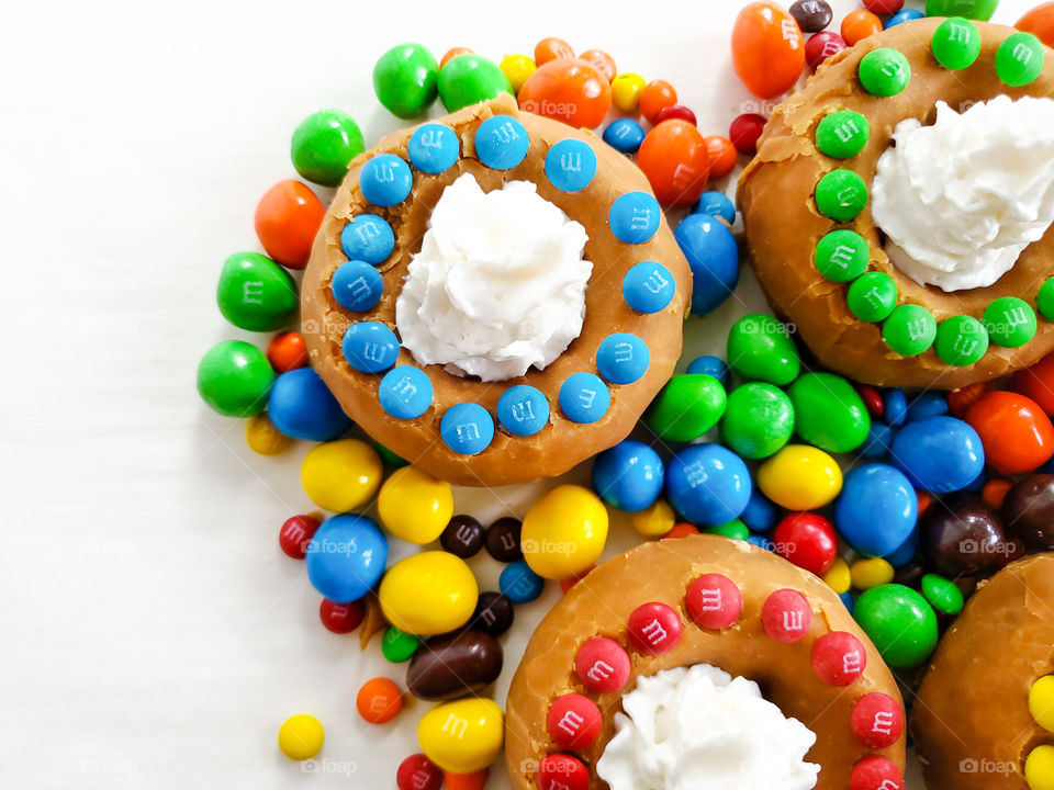 Donuts decorated with M&M minis, topped off with whip cream and surrounded by peanut M&Ms on a white surface.