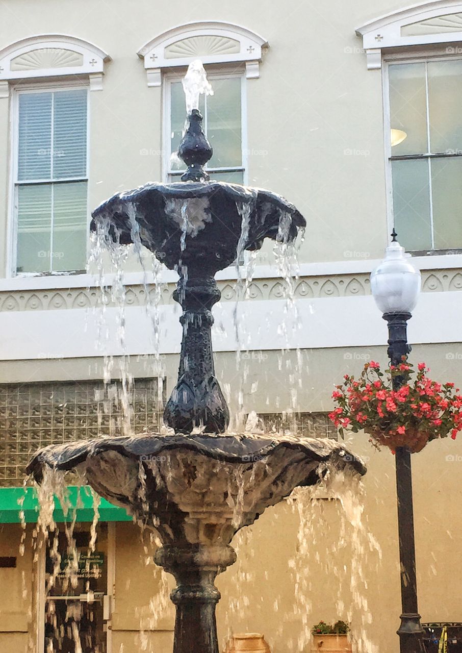 Street scene of city square with a fountain and light post with a hanging basket of flowers