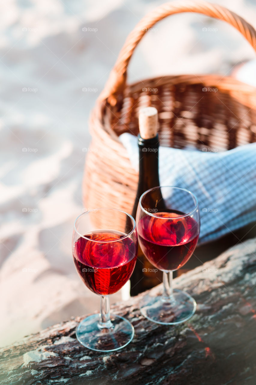 Two wine glasses with red wine standing on tree trunk, on beach, beside wicker basket with bottle of wine