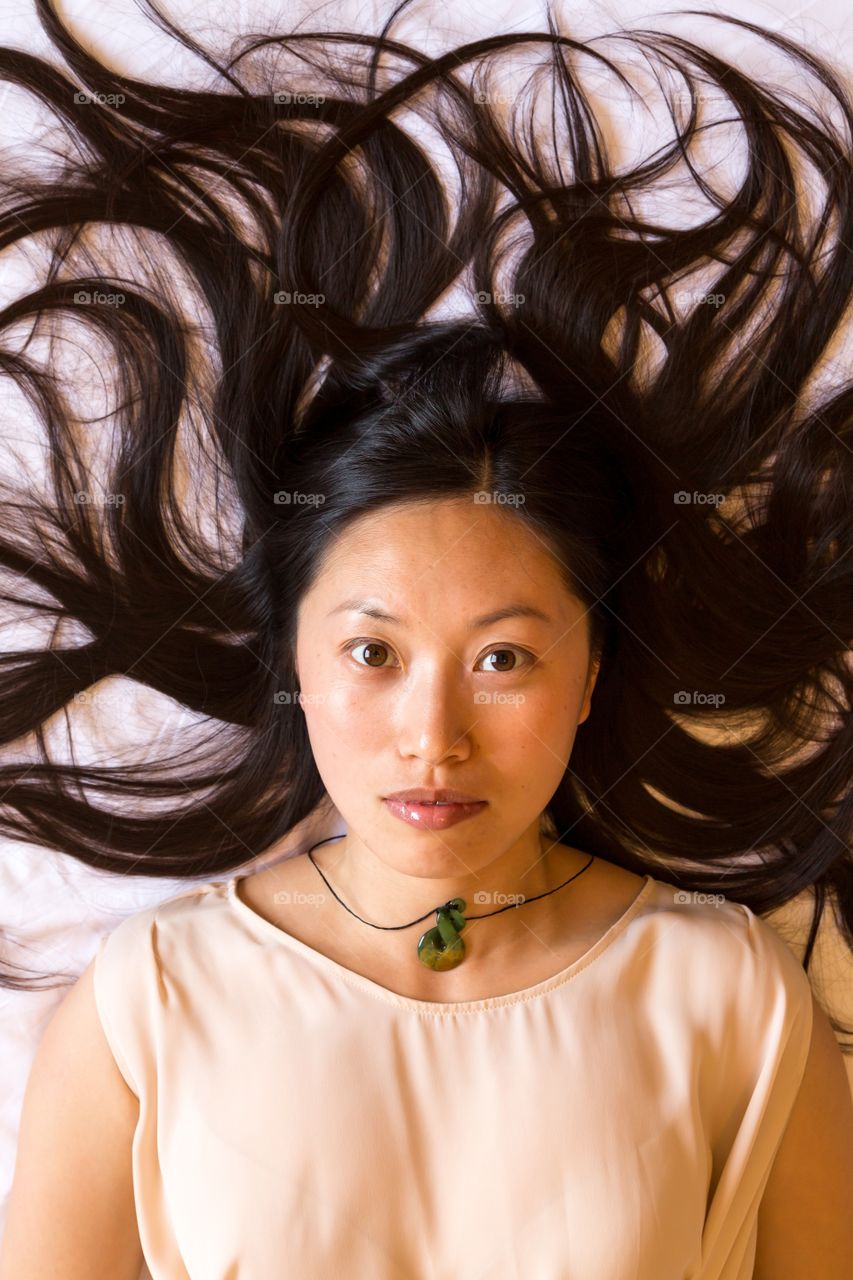 Asian girl posing for camera. Asian girl laying down and looking up towards camera. Hair spread around to fill the frame. Green necklace