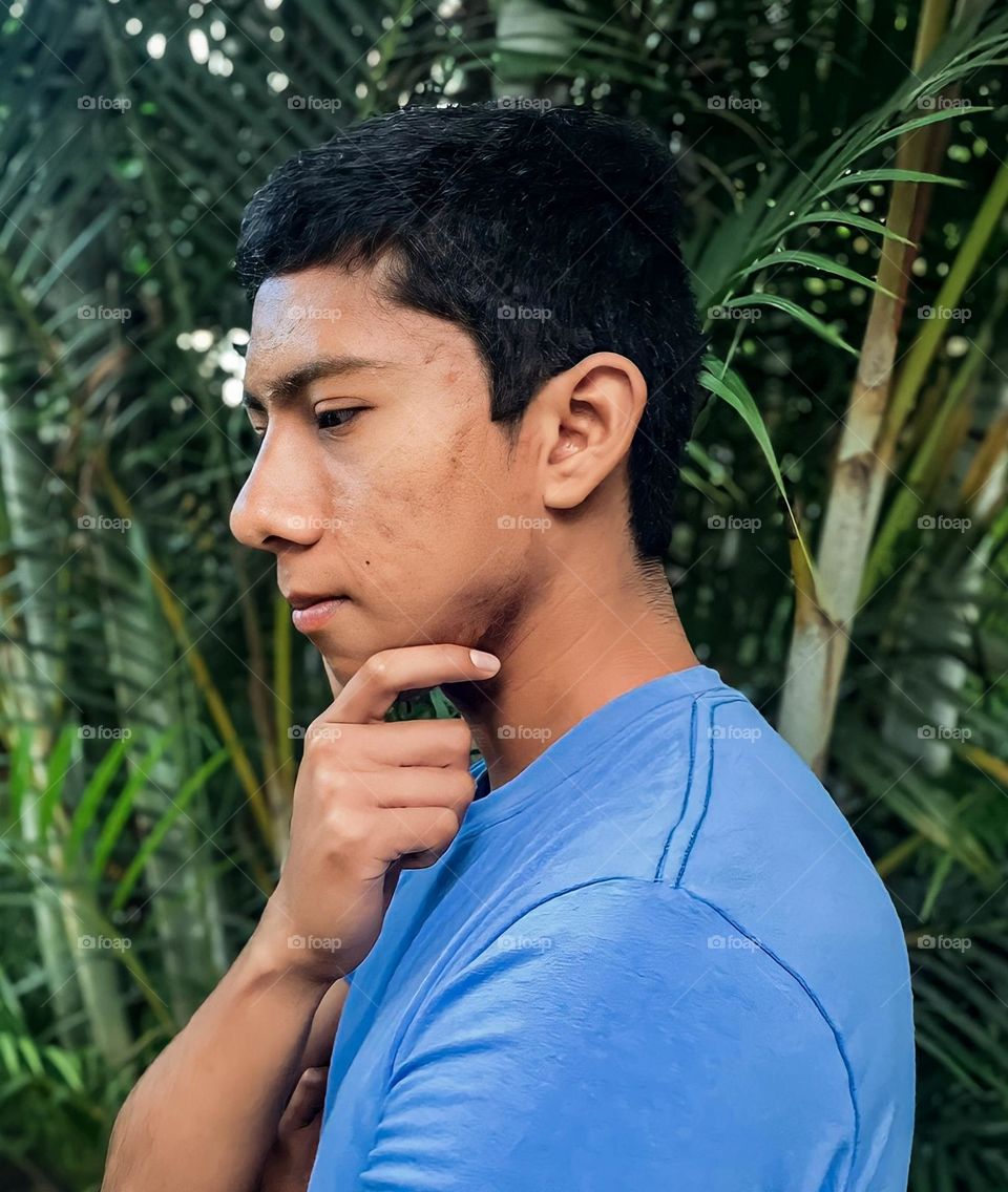 Dark-skinned man, with acne and acne scars, wearing a light blue t-shirt, seen in profile with a thoughtful pose, on a nature background.