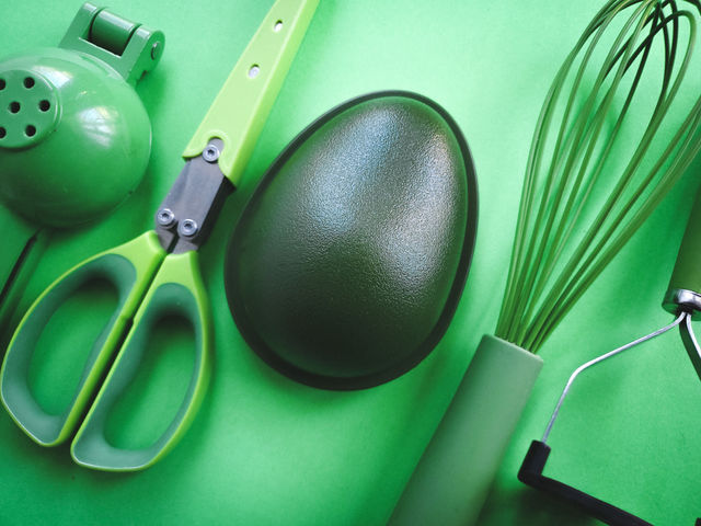 Five green kitchen utensils placed in a diagnol fashion: lemon/lime squeezer/press, herb mincing scissors,  avocado holder container, silicone coated wire wisk, and a masher.