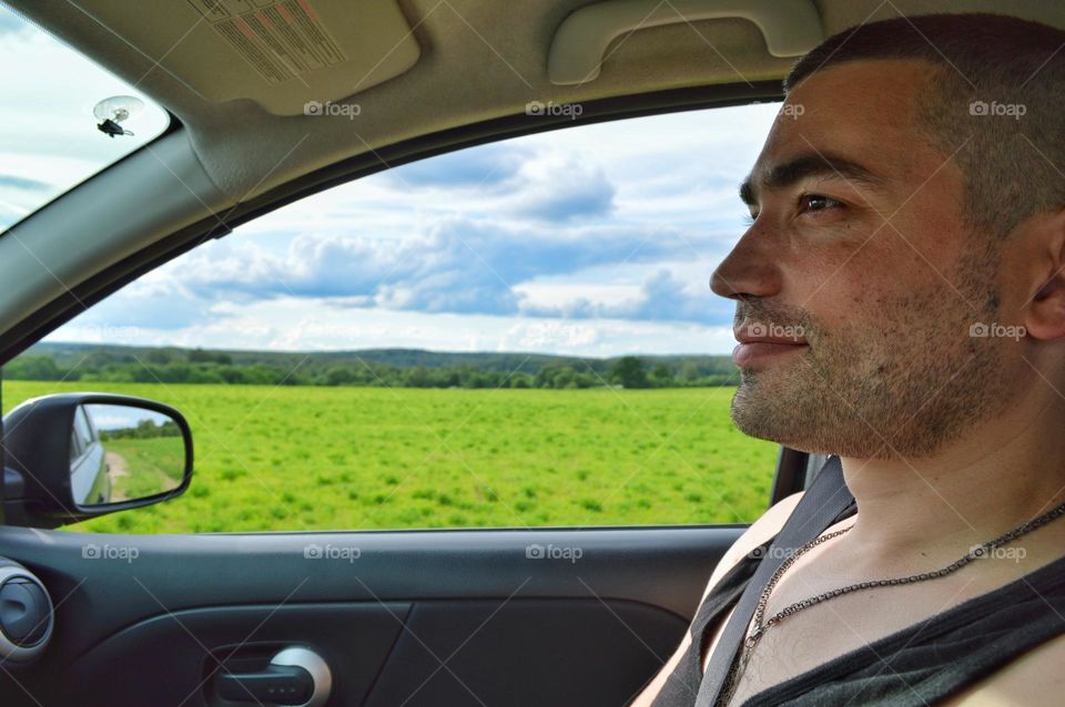 a man in the passenger seat of a car during a trip out of town. outside the window a landscape from a forest field and a cloud