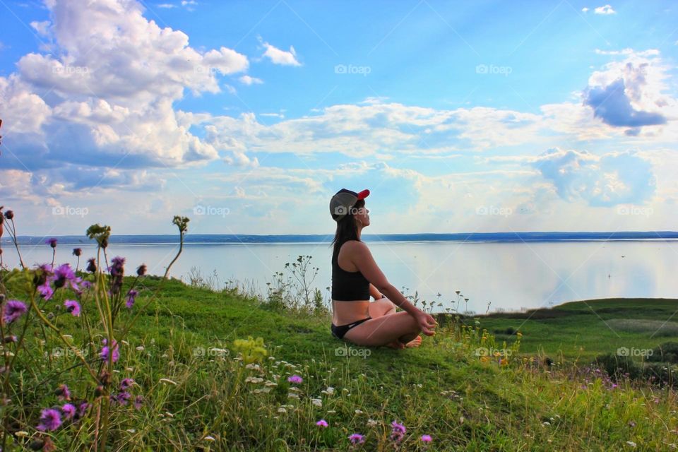 a girl sits in a meditative pose on a hillside in a small clearing against the background of white clouds of a blue sky of a small lake