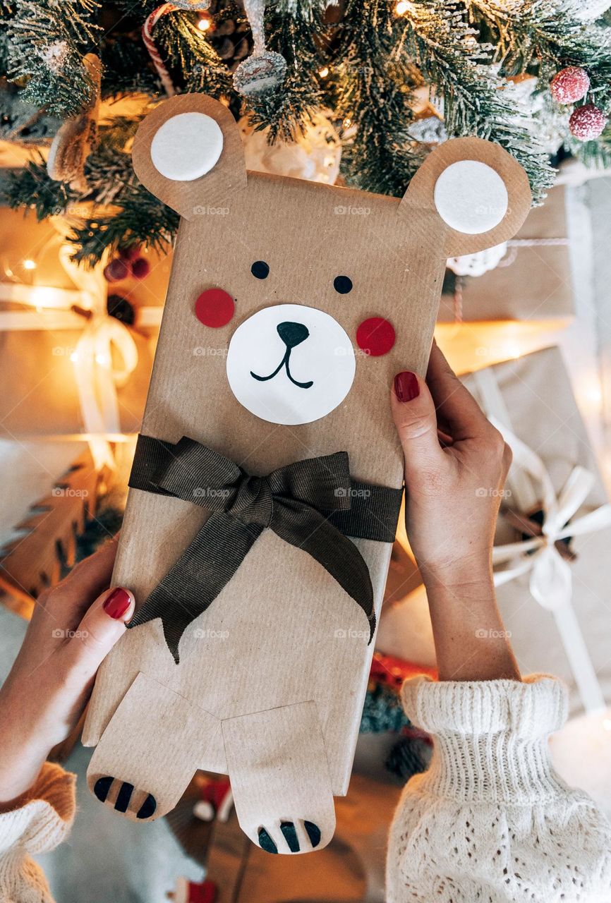 Close-up image of woman's hands holding wrapped christmas present shaped as a teddy bear in front of christmas tree.