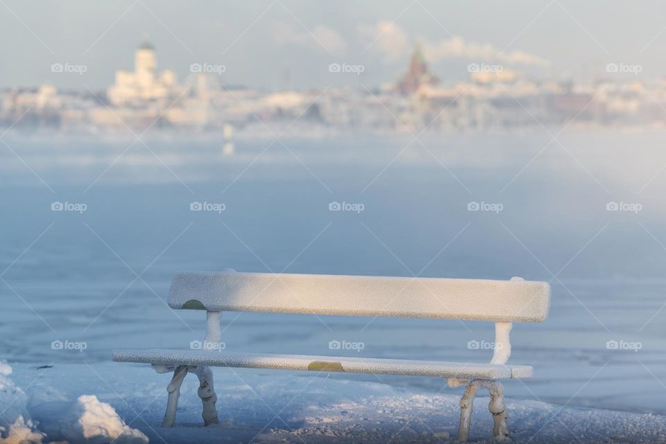 Traditional wooden and frosty resting bench in Helsinki, Finland on an extremely cold winter morning with freezing Baltic Sea and downtown Helsinki skyline in the background.