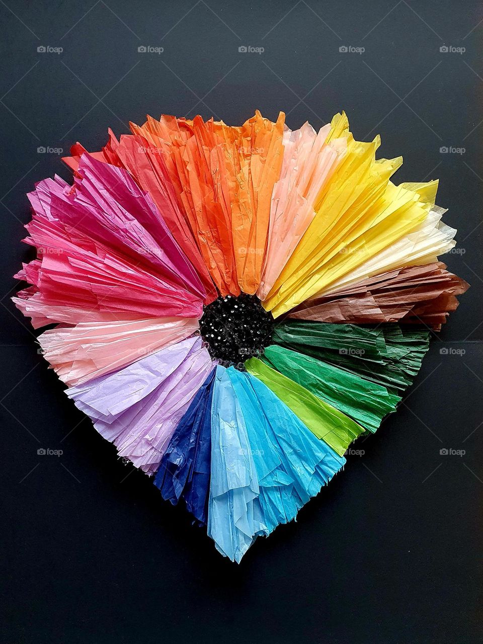 Composition of a heart-shaped flower made of multi-coloured silk papers and black beads, placed on black paper creating a colour explosion