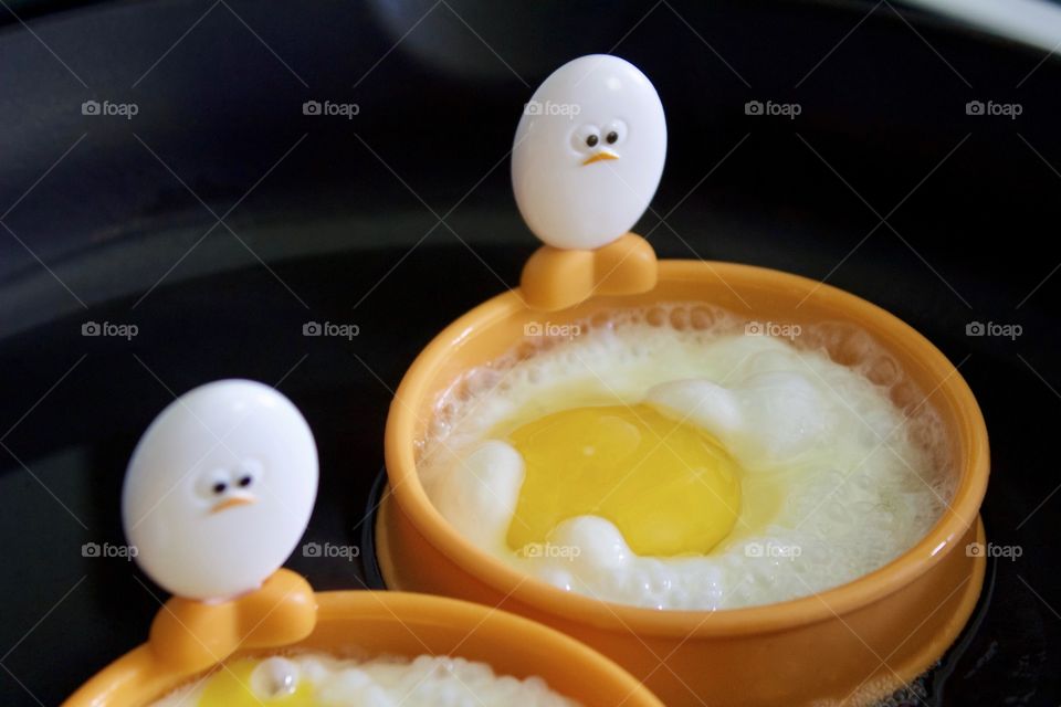 Closeup of a cast iron pan containing eggs being fried in silicone egg rings with egg-shaped handles depicting baby chick faces and feet