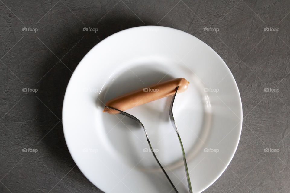 Two forks prick one sausage on a white porcelain plate.