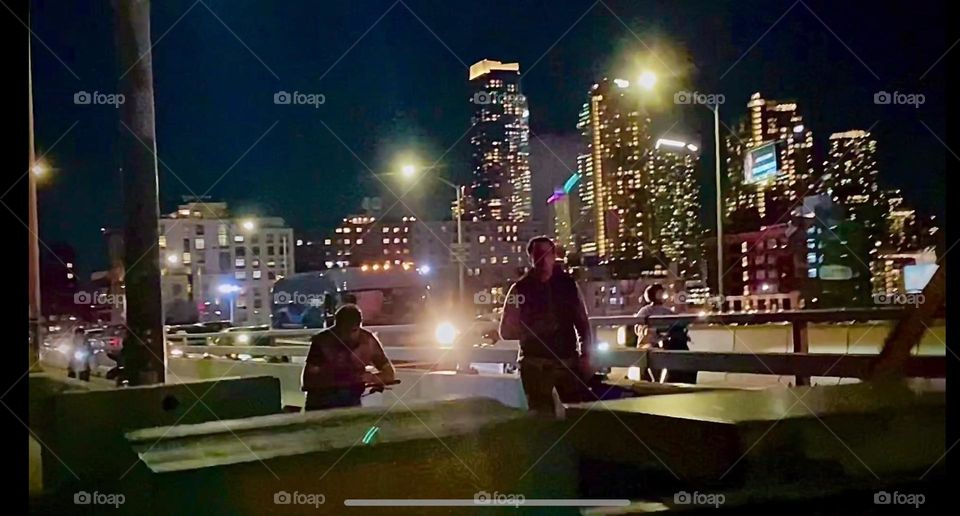 This is the magical view of NYC at night. From the „Pulaski Bridge“ we see the skyline of LIC, Queens sparkling from myriads of electric lights. Two people are waiting for the bridge to reopen for traffic. 2023. Hypnotic Productions