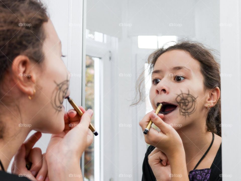 Portrait of a beautiful caucasian teenage girl painting her lips with a black pencil and reflecting in the mirror on the wardrobe in her room, close-up side view.