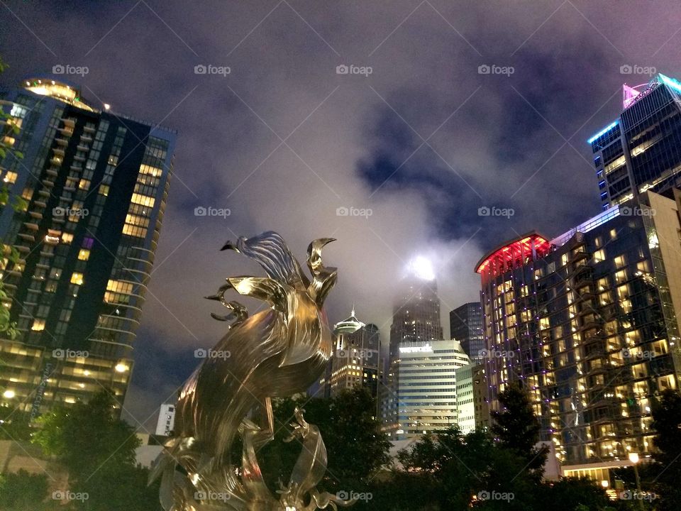 Fantastic beast metal sculpture during dark, cloudy & stormy Uptown Charlotte night skyline with high buildings lit.