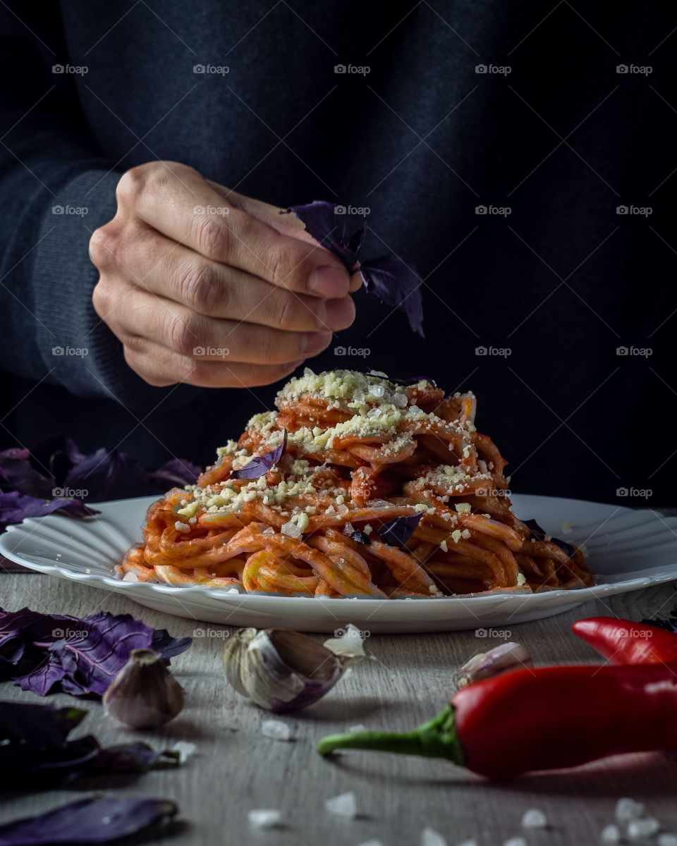 Hot spicy pasta with Diablo sauce. Man is decorating it with putting basil leaf on the top of dish. Ingredients: Italian spaghetti, tomatoes, chili peppers, garlic, sea salt, parmesan cheese, basil.