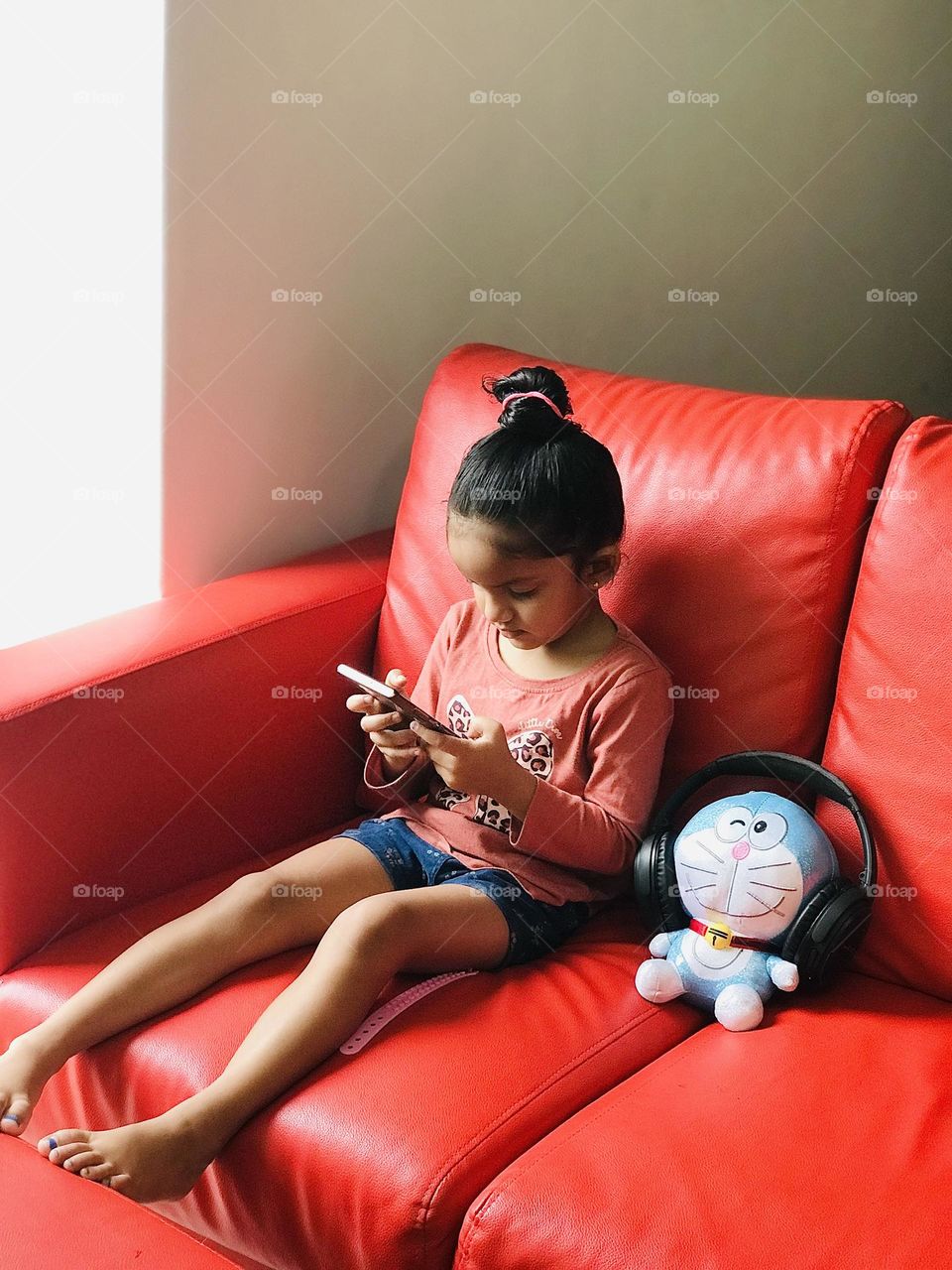 A girl watching phone along with her toy on red couch 