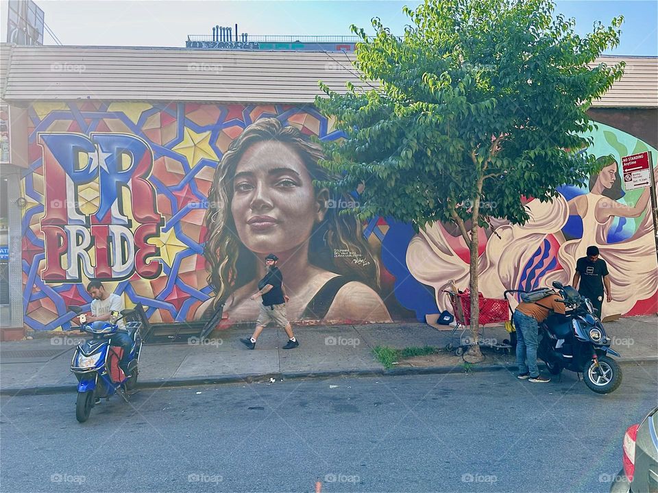 A smiling Latina next to the words “PR” and “PRIDE” on a 1970s style pop art background invites us to celebrate the hispanic culture with this beautiful art mural on “Jefferson St” and “Broadway” in “Bushwick”, Brooklyn. 2024. Hypnotic Productions