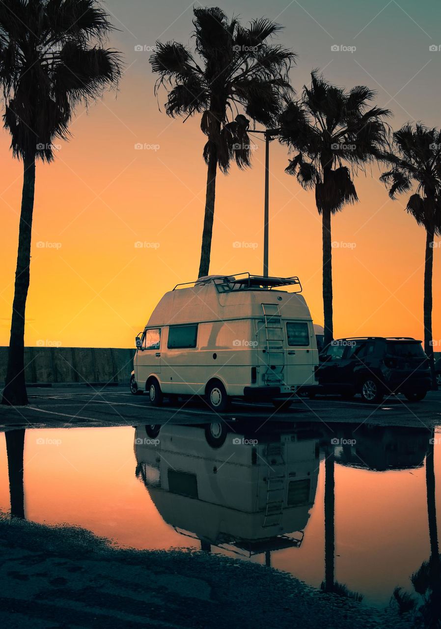 White camping van by the beach at orange sunset under palm trees